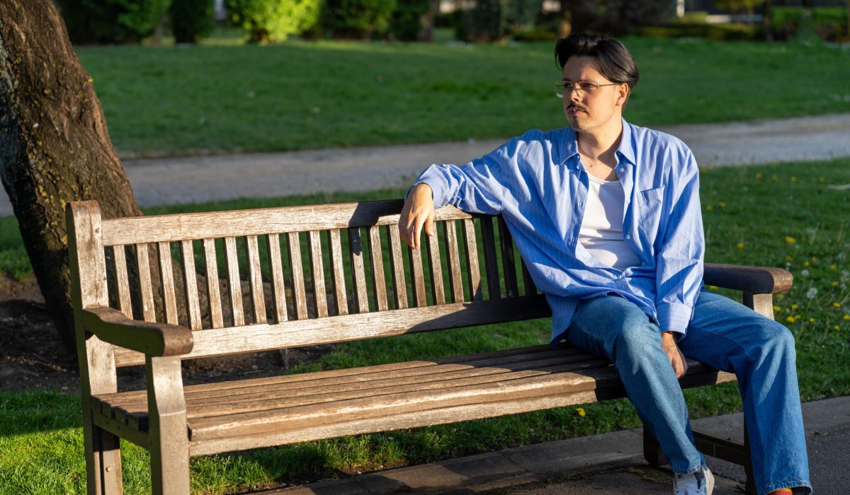 A man in a light blue shirt and jeans sits on a wooden park bench, looking thoughtfully to his left. Sunlight casts a warm glow over him and the surrounding grassy area.