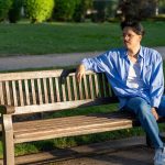 A man in a light blue shirt and jeans sits on a wooden park bench, looking thoughtfully to his left. Sunlight casts a warm glow over him and the surrounding grassy area.