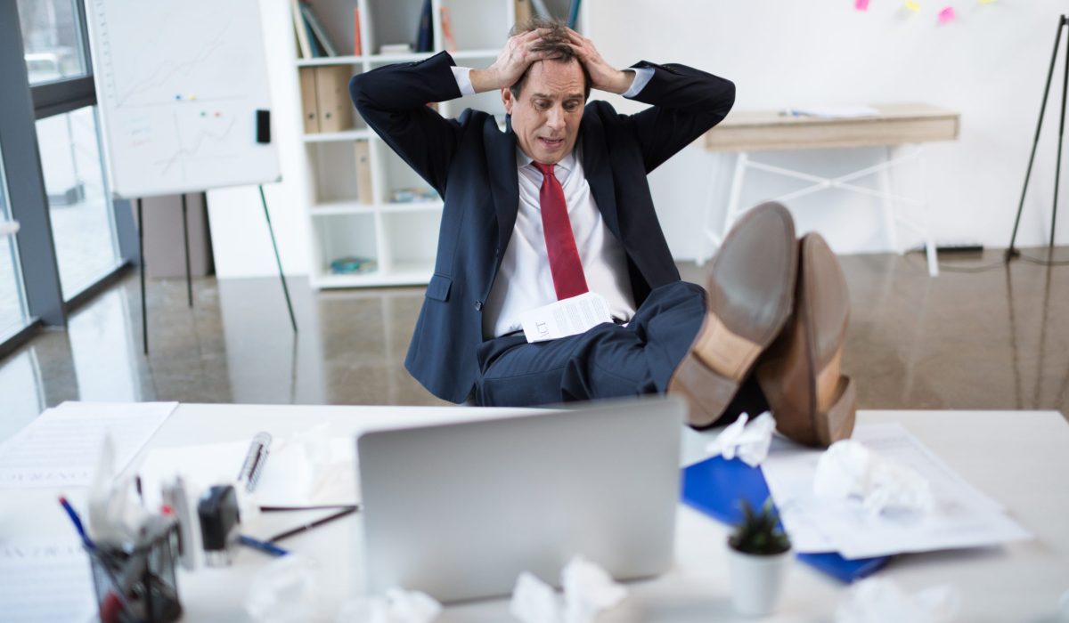 Stressed businessman sitting at his desk with hands on his head, surrounded by crumpled papers.