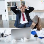 Stressed businessman sitting at his desk with hands on his head, surrounded by crumpled papers.