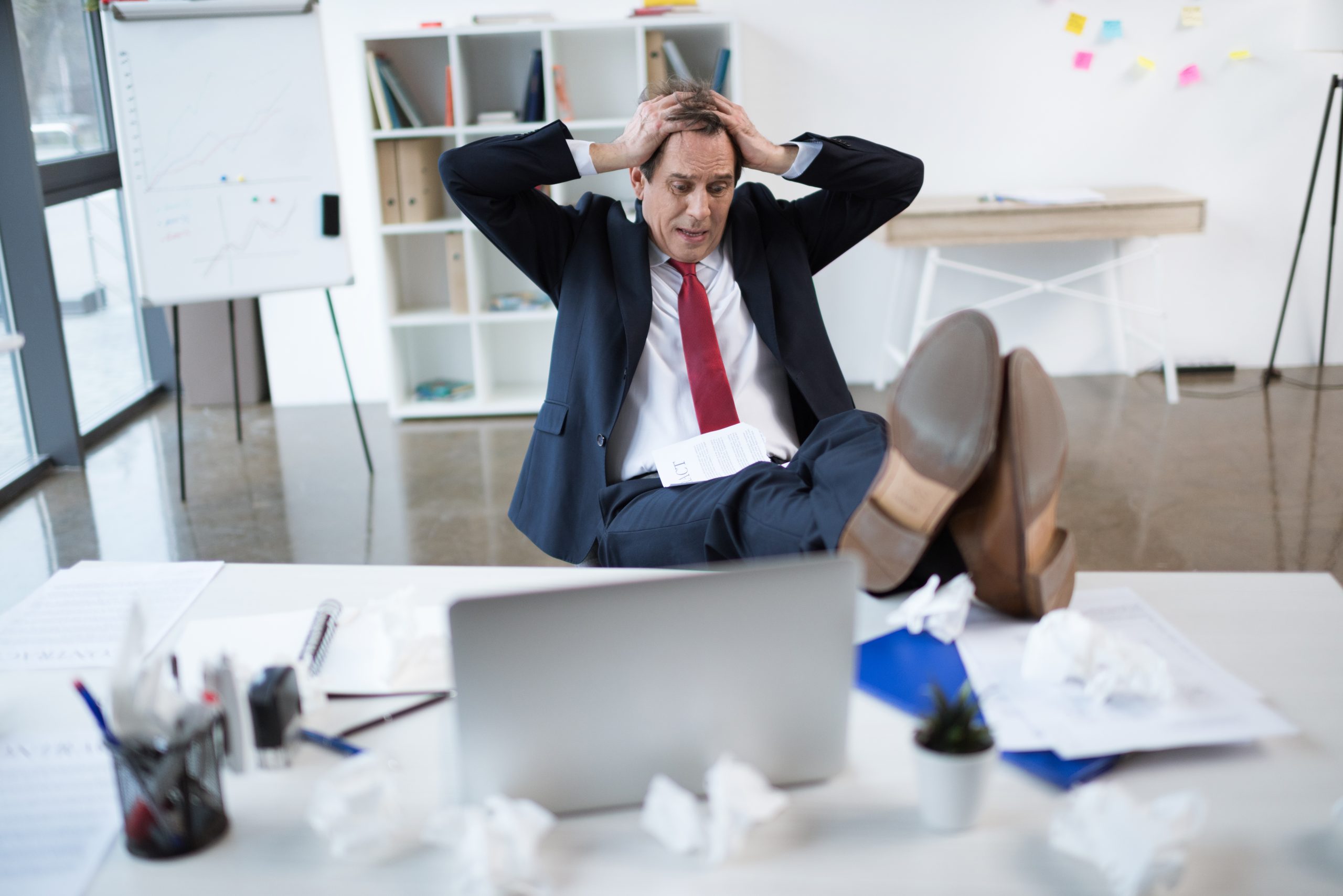 Stressed businessman sitting at his desk with hands on his head, surrounded by crumpled papers.