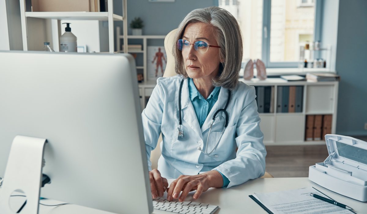 Experienced female doctor in a white lab coat using a computer in her medical office, optimizing patient management with AI technology.